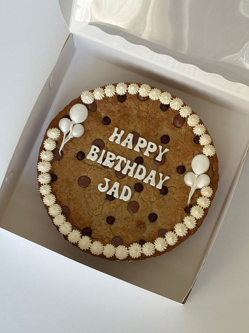 A large chocolate chip cookie decorated with white frosting piped borders, chocolate chips, and 'HAPPY BIRTHDAY JAD' written with edible candy, presented inside a white box.